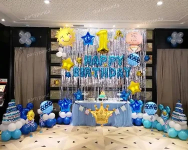 A 1st birthday party decoration setup with a shimmering silver tinsel backdrop, blue "HAPPY BIRTHDAY" foil balloons, a large gold number 1 balloon, and blue and white balloon pillars, featuring a faint "partypanda" watermark.